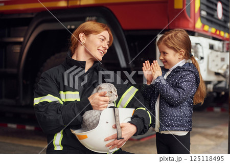 Cat is sitting in the helmet, kid is happy, clapping her hands. Firefighter woman in uniform is with a little girl 125118495