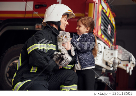Cute scottish fold cat is in the hands. Firefighter woman in uniform is with a little girl Cute scottish fold cat is in the hands. Firefighter woman in uniform is with a little girl 125118503