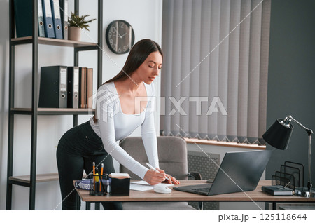 With laptop. Standing and working. Young beautiful woman in white clothes is in the office 125118644