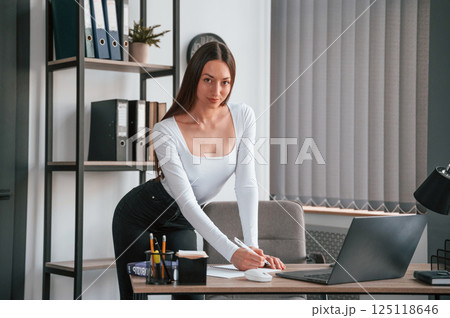 Standing by the table. Young beautiful woman in white clothes is in the office 125118646