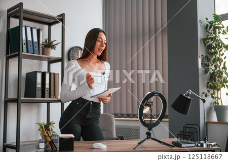 Smartphone is on the stand, recording. Young beautiful woman in white clothes is in the office Smartphone is on the stand, recording. Young beautiful woman in white clothes is in the office 125118667