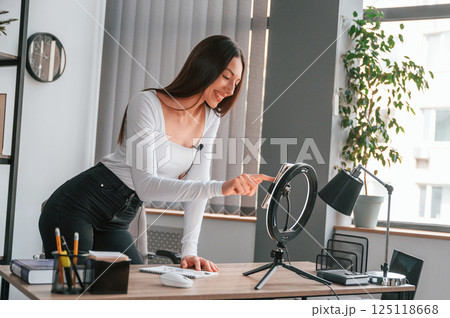 Setting up the smartphone for recording. Young beautiful woman in white clothes is in the office 125118668