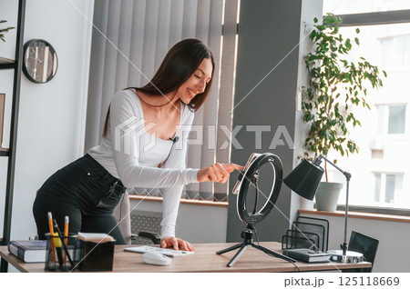 Setting up the smartphone for recording. Young beautiful woman in white clothes is in the office 125118669