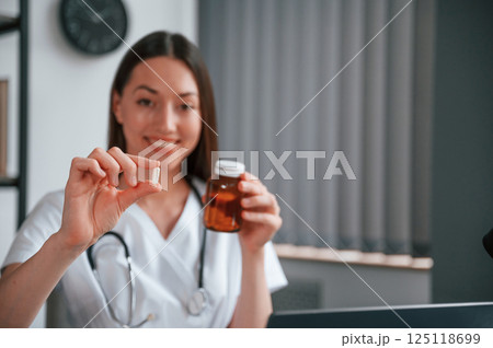Holding pills in hands. Young beautiful female doctor in white coat is in the office Holding pills in hands. Young beautiful female doctor in white coat is in the office 125118699