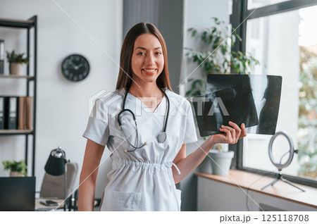 Holding x-ray of bones. Young beautiful female doctor in white coat is in the office Holding x-ray of bones. Young beautiful female doctor in white coat is in the office 125118708