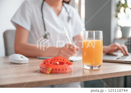 Close up view. Apple juice and measuring length is on the table. Young beautiful female doctor with laptop 125118744