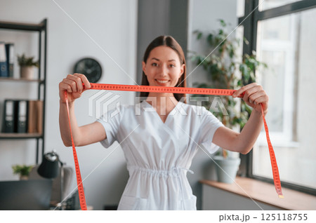 Standing with measuring tape. Young beautiful woman in white clothes is in the office Standing with measuring tape. Young beautiful woman in white clothes is in the office 125118755