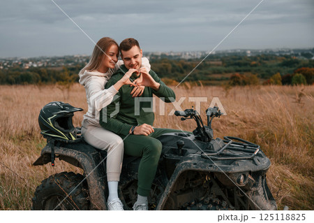 Making love shape gesture by hands. Beautiful young couple is with quad bike outdoors on the field 125118825