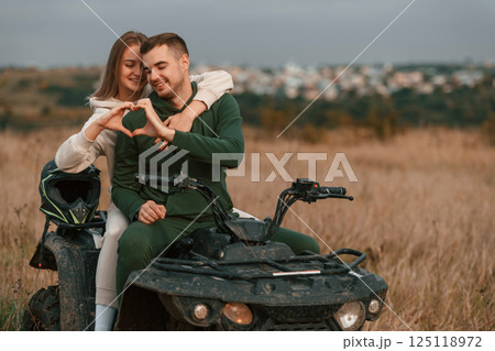 Making love shape gesture by hands. Beautiful young couple is with quad bike outdoors on the field Making love shape gesture by hands. Beautiful young couple is with quad bike outdoors on the field 125118972