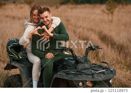 Making love shape gesture by hands. Beautiful young couple is with quad bike outdoors on the field Making love shape gesture by hands. Beautiful young couple is with quad bike outdoors on the field 125118973