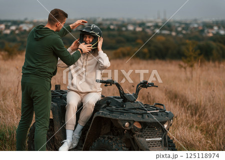 Man is helping his girlfriend to wear a helmet. Lovely young couple is with quad bike outdoors on the field 125118974