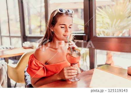 woman in a cafe seated, holding beverage. Dressed in an orange dress with her hair down. Bright interior, large windows allow natural light. Beverage consumption for refreshment. 125119143