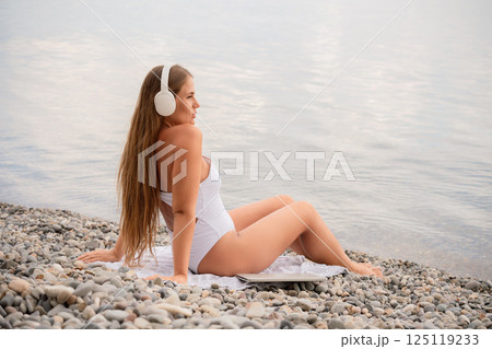 A woman is sitting on a beach with her headphones on. She is wearing a white bikini and a white shirt. The beach is rocky and the water is calm. The woman is enjoying her time by the water. 125119233
