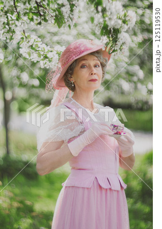 Natural adult mature woman of 60 years old in 18th century style pink hat, retro dress, gloves holds cup of tea in blossoming white apple orchard on summer day Natural adult mature woman of 60 years old in 18th century style pink hat, retro dress, gloves holds cup of tea in blossoming white apple orchard on summer day 125119530
