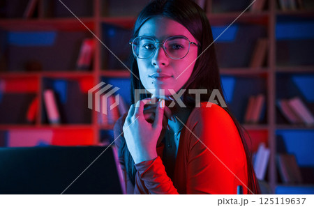 Beautiful woman in glasses and red wear is sitting by the laptop in dark room with neon lighting 125119637