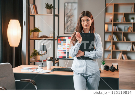 Standing against pc and holding notepad. Young female designer is in the modern office Standing against pc and holding notepad. Young female designer is in the modern office 125119814