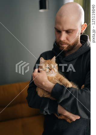 Vertical portrait of serious bearded bald man wearing dark hoodie, gently holding ginger cat in arms, creating heartwarming scene of companionship and love within cozy home environment. Vertical portrait of serious bearded bald man wearing dark hoodie, gently holding ginger cat in arms, creating heartwarming scene of companionship and love within cozy home environment. 125119840