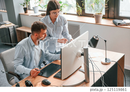 Man is sitting, woman is standing. Using pc. Two young designers is working together in the office 125119861