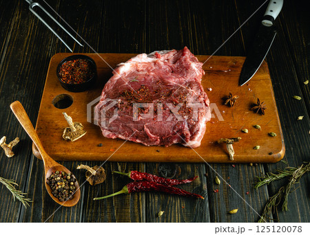 A generous cut of raw beef is placed on a wooden cutting board, surrounded by an array of spices. The rich marbling of the meat is highlighted as it awaits seasoning for a delicious meal 125120078