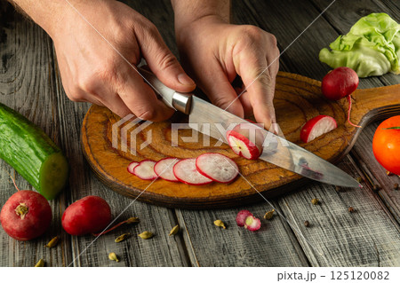 Hands skillfully slice vibrant radishes on a rustic wooden cutting board surrounded by an array of fresh vegetables including cucumbers and lettuce. Sunlight illuminates the work area 125120082