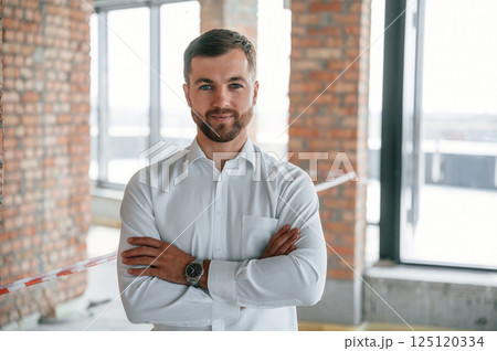 Standing with arms crossed. Man in white shirt is working indoors at construction site. Managing the project Standing with arms crossed. Man in white shirt is working indoors at construction site. Managing the project 125120334