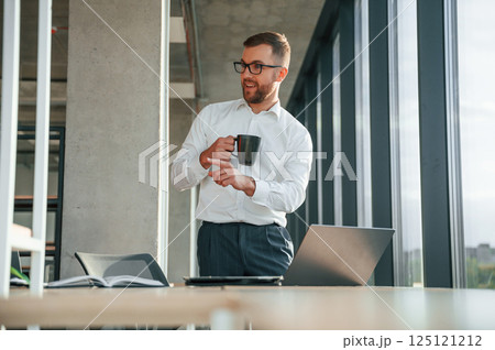 Holding cup of drink. Man in formal clothes is working in the modern office 125121212