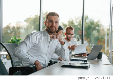 Man is looking at the camera. Three people in formal clothes are working in the modern office together 125121231