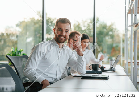 Man is looking at the camera. Three people in formal clothes are working in the modern office together 125121232