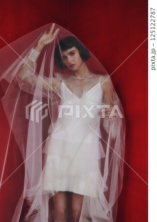 Elegant model poses in sheer fabric dress against a vibrant red background during a fashion photoshoot Elegant model poses in sheer fabric dress against a vibrant red background during a fashion photoshoot 125122787