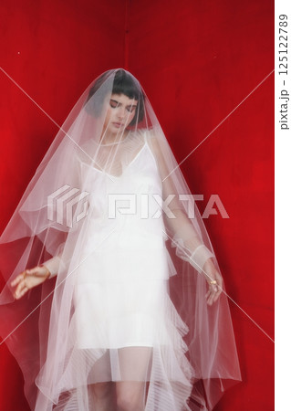 Bride in a white dress and sheer veil poses gracefully against a vibrant red backdrop Bride in a white dress and sheer veil poses gracefully against a vibrant red backdrop 125122789