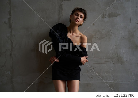 Stylish model poses in elegant black outfit against textured gray wall in indoor setting 125122790