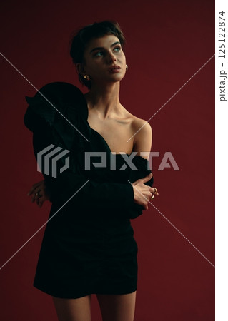Stylish model poses in a black dress against a deep red backdrop during a studio photoshoot Stylish model poses in a black dress against a deep red backdrop during a studio photoshoot 125122874