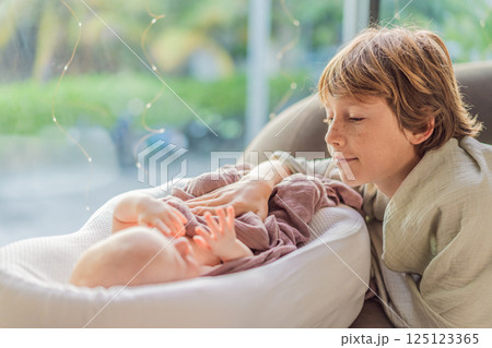 Older brother spending quality time with his newborn sibling as the baby rests in a cozy cocoon. Family bonding, love, and childhood connection concept 125123365