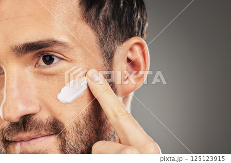 Face, skincare and beauty with a man model using treatment in studio on a gray background for wellness. Finger, cosmetics and antiaging with a handsome young male inside to apply lotion to his cheek 125123915