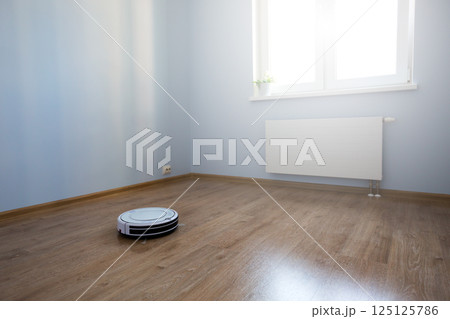 White robot vacuum cleaner on laminate floor cleaning dust in empty room interior White robot vacuum cleaner on laminate floor cleaning dust in empty room interior 125125786