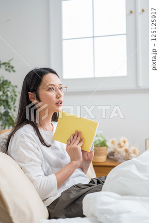 室内で手帳を見て考えるメガネ女性｜日記読書｜健康的な暮らしイメージ｜ウェルビー女子｜ウェルビーイング 125125917