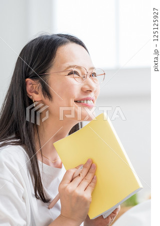 室内で手帳を見る笑顔のメガネ女性｜日記読書｜健康的な暮らしイメージ｜ウェルビー女子｜ウェルビーイング 125125927
