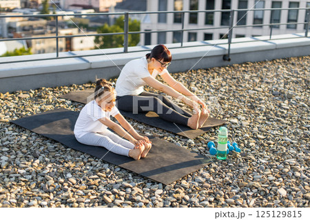 Mother and daughter practicing stretching exercises outdoors on rooftop during sunny day. Fitness mat and water bottle showing workout essentials. 125129815