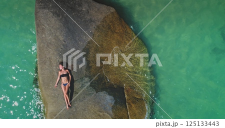 A person enjoys the warmth of the sun while reclining on a large rock, surrounded by clear turquoise water and a serene atmosphere. The bright summer day creates a perfect relaxing getaway. 125133443