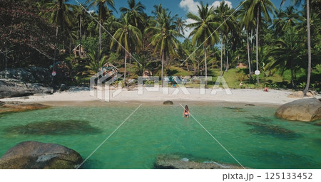 Crystal clear water gently laps at the shore as a person stands in the shallow part of the beach. Lush palm trees surround the tranquil scene, indicating a warm, sunny day. Crystal clear water gently laps at the shore as a person stands in the shallow part of the beach. Lush palm trees surround the tranquil scene, indicating a warm, sunny day. 125133452