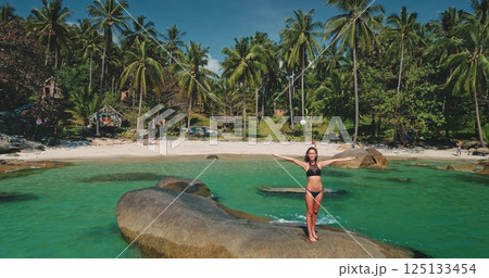 A woman stands on a rock, arms outstretched, at a picturesque beach surrounded by palm trees and clear turquoise water. It's a sunny day, perfect for relaxation and fun. A woman stands on a rock, arms outstretched, at a picturesque beach surrounded by palm trees and clear turquoise water. It's a sunny day, perfect for relaxation and fun. 125133454