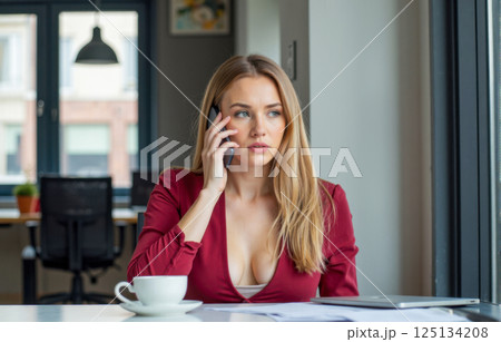 Attractive young woman in red outfit talking on phone, sitting in modern office, holding smartphone, looking away, coffee cup, natural light, thoughtful expression 125134208