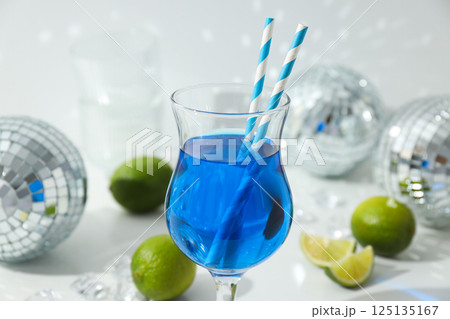 Blue cocktail in glass, limes, ice cubes and disco balls on white background, close up Blue cocktail in glass, limes, ice cubes and disco balls on white background, close up 125135167