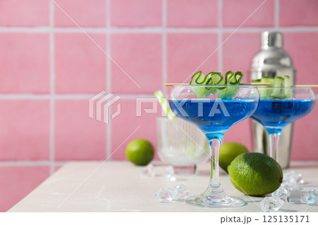 Blue cocktail with cucumber slice in glasses, shaker, limes and ice cubes on pink background, space for text 125135171