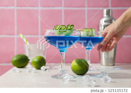 Blue cocktail with cucumber slice in glasses, hand, shaker, limes and ice cubes on pink background Blue cocktail with cucumber slice in glasses, hand, shaker, limes and ice cubes on pink background 125135172