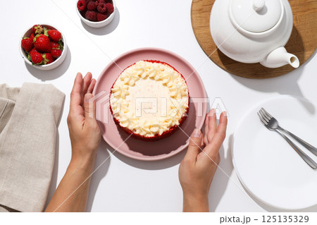 Red velvet cake on plate in female hands, bowls with berries and teapot on white background, top view Red velvet cake on plate in female hands, bowls with berries and teapot on white background, top view 125135329