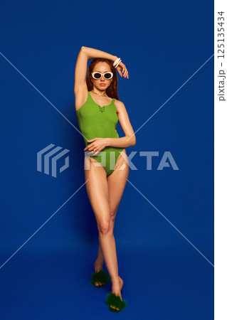 Elegant model in green swimsuit and fluffy heels standing with crossed legs and arm lifted in graceful pose against blue studio background. 125135434