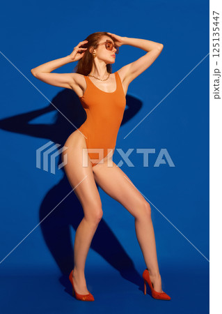 Elegant young model striking fashion pose in orange swimsuit and heels with shadow dancing behind her against blue studio background. Elegant young model striking fashion pose in orange swimsuit and heels with shadow dancing behind her against blue studio background. 125135447