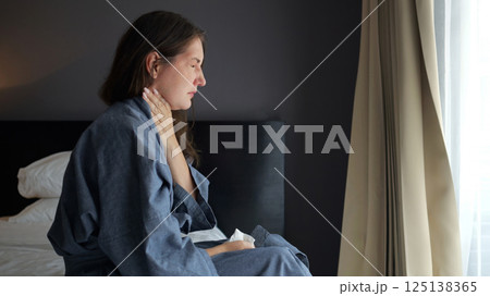 Young caucasian woman in bathrobe sitting on bed holding throat with painful expression 125138365