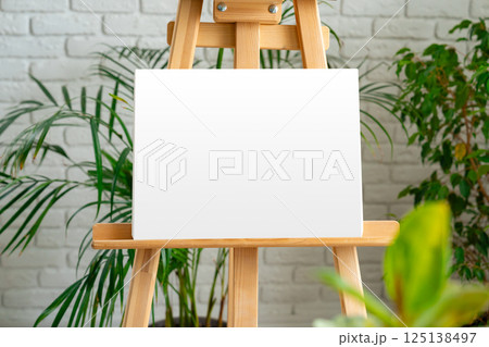 Blank canvas on an easel surrounded by greenery in a bright, modern art studio 125138497